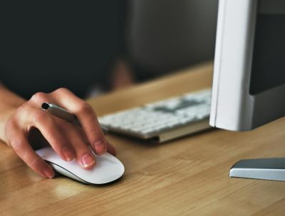 A hand using a wireless mouse at a modern desk setup with a computer and keyboard.